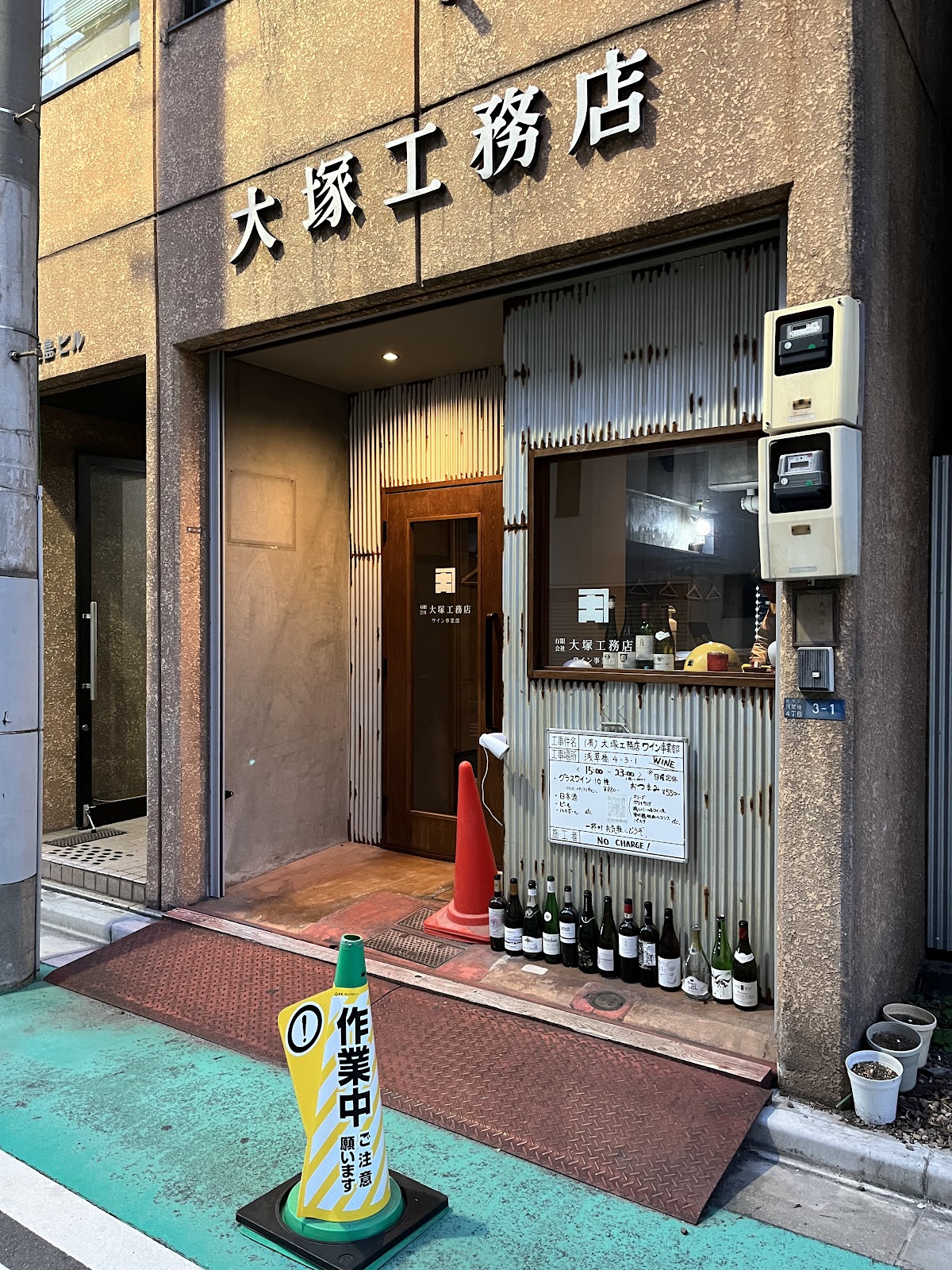 昼の店舗外観。古いビルの一階、シャッター風の鉄扉と工事用パイロン